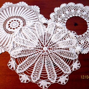 May include: Three white, handmade crochet doilies with intricate lace patterns. The doilies are circular, with varying designs including a central floral motif and scalloped edges. The date "12/04/2025" is visible in the lower right corner.