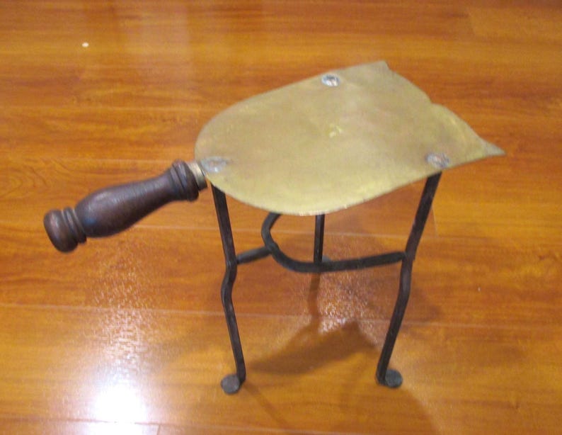 May include: An antique brass trivet with a dark wooden handle and a black metal stand. The trivet has a heart-shaped brass top and is designed to protect surfaces from hot cookware. The handle is attached with a brass fitting.