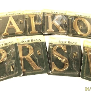 May include: A collection of solid brass letters, including A, H, K, O, P, R, S, and W, displayed in their original packaging. Each letter is a warm gold color and comes with mounting screws. The packaging reads "SOLID BRASS".
