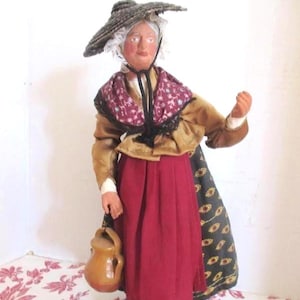 May include: A handcrafted figurine of a woman in traditional attire. She wears a wide-brimmed hat, a burgundy skirt, and a patterned shawl. The figure holds a brown jug and is posed as if walking.