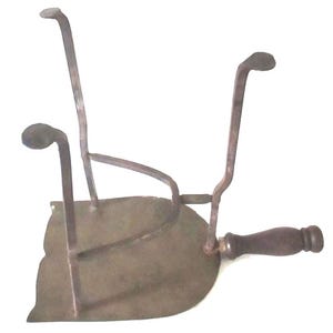 May include: Antique metal iron rest with a dark wooden handle. The iron rest has a flat, curved base with three upright supports. The metal has a weathered, aged appearance, suggesting it is an antique or vintage item.