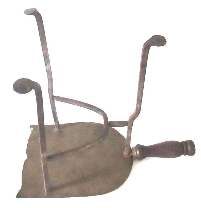 May include: Antique metal iron rest with a dark wooden handle. The iron rest has a flat, curved base with three upright supports. The metal has a weathered, aged appearance, suggesting it is an antique or vintage item.