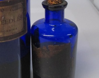 May include: Two cylindrical cobalt blue glass apothecary bottles with narrow necks and cork stoppers. The smaller bottle features a dark, weathered paper label. The larger bottle has a partially visible label with the text NEEDLES and ROBBINS.