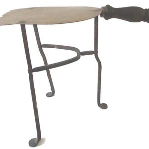 May include: An antique trivet with a metal frame and a wooden handle. The trivet has a three-legged base and a flat, leaf-shaped top. The handle is dark brown, and the metal frame is a dark, aged color.
