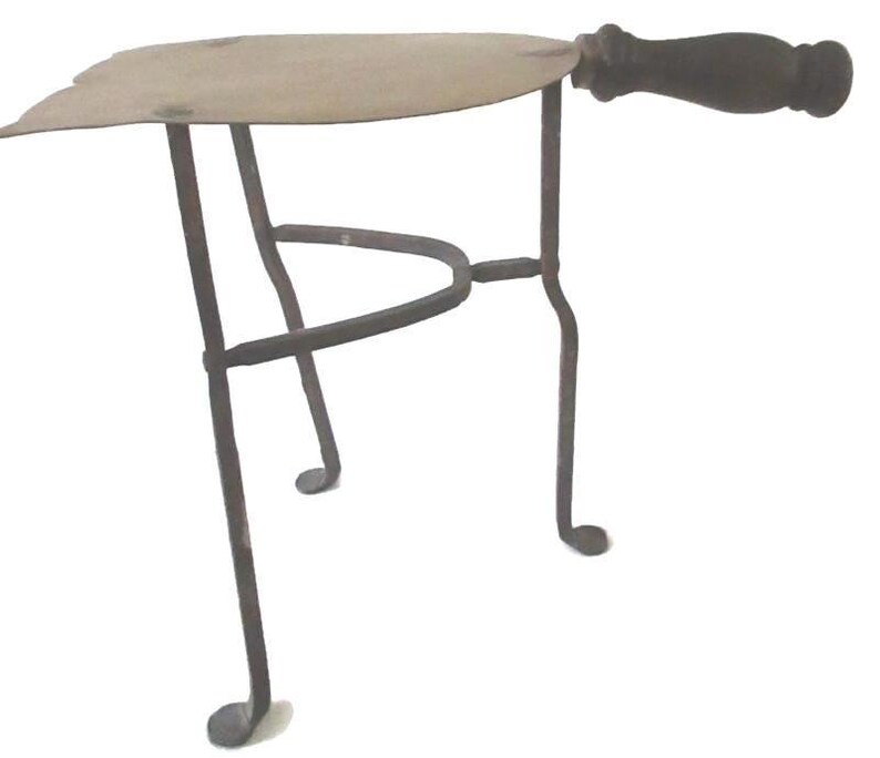 May include: An antique trivet with a metal frame and a wooden handle. The trivet has a three-legged base and a flat, leaf-shaped top. The handle is dark brown, and the metal frame is a dark, aged color.