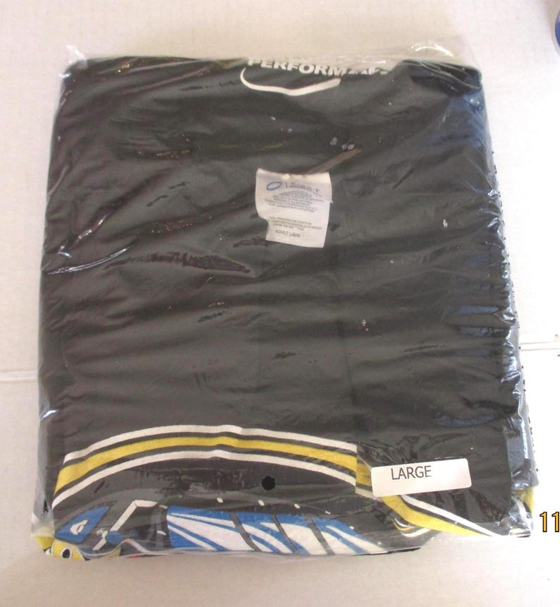May include: A black t-shirt sealed in a clear plastic bag. The shirt features a design with yellow, white, and blue details. A label marked "LARGE" is affixed to the packaging.