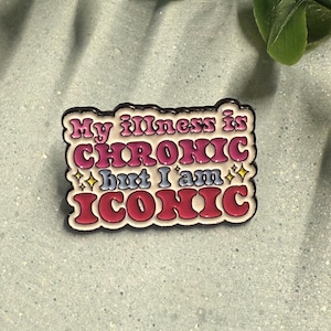 Op de afbeelding: Een rechthoekige pin met de tekst "My illness is CHRONIC but I am ICONIC" in roze, blauwe en gele letters. De pin heeft een zwarte rand en staat op een lichtblauwe achtergrond met groen blad.