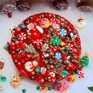 Könnte beinhalten: Ein kreisförmiges rotes Harzstück mit eingebetteten weihnachtlichen Anhängern wie Weihnachtsmann, Rentieren, Schneemännern, Tannenbäumen und Zuckerstangen, umgeben von losen Weihnachtsanhängern, Tannenzapfen und einer Miniatur-Eisenbahn.