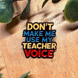 Op de afbeelding: Een rechthoekige speld met de tekst "DON'T MAKE ME USE MY TEACHER VOICE" in geel, blauw en rood. De speld is geplaatst tegen een houten achtergrond, omringd door groene bladeren.