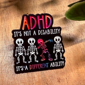 Pode incluir: Um adesivo preto para TDAH com o texto "ADHD It's Not A Disability It's A Different Ability". O adesivo apresenta quatro figuras de esqueleto, uma delas fazendo um dab em tons coloridos. A palavra TDAH está em cores de arco-íris.
