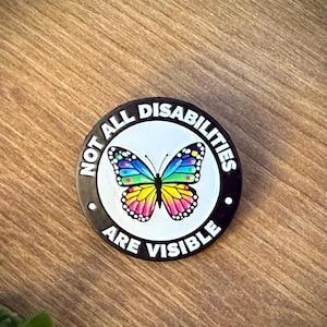 Peut inclure: Un pin's rond en émail avec un centre blanc et une illustration de papillon aux couleurs de l'arc-en-ciel. La bordure noire extérieure porte le texte blanc "NOT ALL DISABILITIES ARE VISIBLE" en lettres capitales grasses.