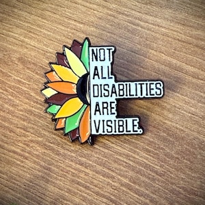Peut inclure: Une broche en émail métallique représentant un tournesol sur le côté gauche avec des pétales jaunes, orange et verts. Le côté droit est une forme rectangulaire noire avec le texte blanc : NOT ALL DISABILITIES ARE VISIBLE. Contour en métal noir.