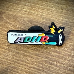 Könnte beinhalten: Ein rechteckiger Emaille-Pin mit schwarzem Rand und weißem Hintergrund, der den Text "POWERED BY ADHD" in bunten Buchstaben zeigt. Das Design enthält eine stilisierte Batterie-Grafik mit gelben Blitzsymbolen auf der rechten Seite.
