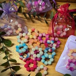 Peut inclure: Plusieurs bagues fleuries en perles colorées, dans des tons de rose, orange, jaune et bleu. Certaines bagues sont en vrac, d'autres dans de petits sacs en organza. Quelques-unes sont dans un petit sac zippé transparent. Fond en bois.