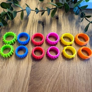 Może przedstawiać: Kolekcja dziesięciu kolorowych pierścieni w różnych odcieniach zieleni, niebieskiego, czerwieni, różu, żółtego i pomarańczowego. Pierścienie mają teksturowane wzory i są ułożone na drewnianej powierzchni, z zielonymi liśćmi w tle.