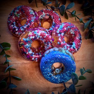 Könnte beinhalten: Fünf bunte Harz-Donuts mit Perlenverzierungen. Die Donuts weisen eine Vielzahl von Farben auf, darunter Rosa, Blau und Lila. Die Donuts sind auf einer Holzoberfläche angeordnet, mit grünem Laub im Hintergrund.