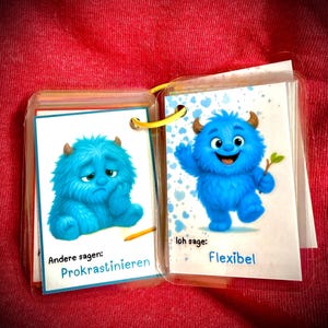 Könnte beinhalten: Ein kleines, offenes Buch mit zwei Seiten, die blaue Cartoon-Monster zeigen. Eines der Monster ist traurig, das andere fröhlich. Auf den Seiten steht "Prokrastinieren" und "Flexibel". Das Buch ist mit einem gelben Ring gebunden.