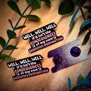 Puede incluir: Tres pines de esmalte con el texto "WELL, WELL, WELL if it isn't the CONSEQUENCES of my own EXECUTIVE DYSFUNCTION" en blanco y rosa sobre fondo negro. Los pines est&aacute;n sobre una superficie de madera con hojas verdes.