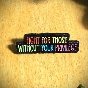 Puede incluir: Un pin rectangular de esmalte negro con bordes redondeados que presenta el texto multicolor "FIGHT FOR THOSE WITHOUT YOUR PRIVILEGE" en una fuente sans-serif. Las letras muestran degradados en tonos naranja, amarillo, verde, azul y rosa.