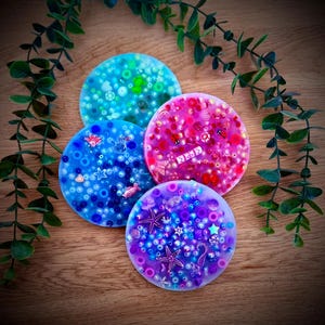 Op de afbeelding: Vier kleurrijke, ronde onderzetters met ingebedde decoratieve elementen. De onderzetters zijn in tinten blauw, roze en paars, met kralen en kleine versieringen in verschillende vormen en maten. Sommige onderzetters hebben zeesterren en zeepaardjes.