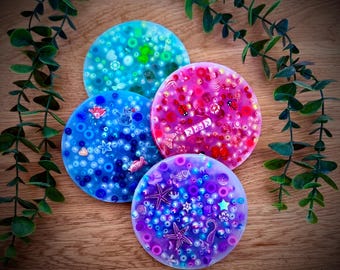 Puede incluir: Cuatro posavasos redondos y coloridos con elementos decorativos incrustados. Los posavasos son en tonos de azul, rosa y morado, con cuentas y pequeños adornos de varias formas y tamaños. Algunos posavasos presentan estrellas de mar e hipocampos.