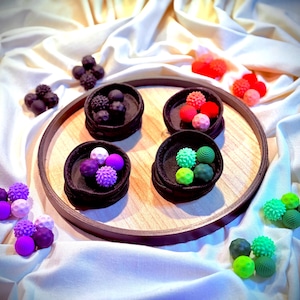 Może przedstawiać: Widok z góry na drewnianą tacę z czterema małymi, ciemnymi, tkanymi koszami, każdy wypełniony kolorowymi, teksturowanymi kulami. Różne kule w odcieniach fioletu, czerwieni i zieleni są rozrzucone wokół tacy na białej tkaninie.