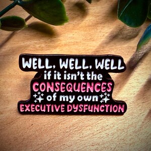 Puede incluir: Pin esmaltado negro con el texto "WELL, WELL, WELL" en blanco, seguido de "if it isn't the CONSEQUENCES of my own EXECUTIVE DYSFUNCTION" en rosa y blanco. El pin est&aacute; sobre una superficie de madera.