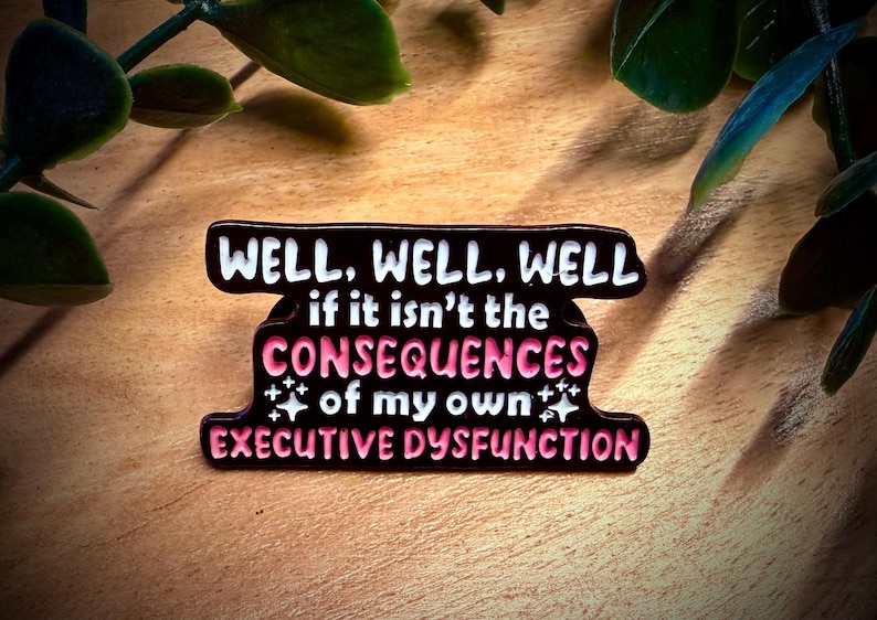 Puede incluir: Pin esmaltado negro con el texto "WELL, WELL, WELL" en blanco, seguido de "if it isn't the CONSEQUENCES of my own EXECUTIVE DYSFUNCTION" en rosa y blanco. El pin est&aacute; sobre una superficie de madera.