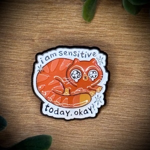 Könnte beinhalten: Eine Emaille-Anstecknadel mit einem orangefarbenen getigerten Kater mit großen, tränenden Augen. Die Katze ist zusammengerollt und der Text "i am sensitive today, okay!" steht in schwarzen Buchstaben um die Katze herum. Der Pin hat einen weißen Hintergrund.