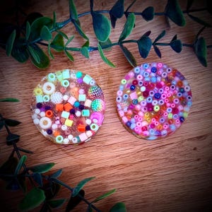Op de afbeelding: Twee ronde hars onderzetters gevuld met kleurrijke kralen en kleine decoratieve elementen. Eén onderzetter heeft oranje, groene en witte kralen, de andere roze en paarse. Ze liggen op een houten oppervlak.