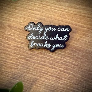 Könnte beinhalten: Eine schwarze Emaille-Anstecknadel in unregelmäßiger, wolkenartiger Form mit dem weißen Schreibschrift-Text Only you can decide what breaks you. Die Anstecknadel hat eine glatte, dunkle Oberfläche mit klarer, gut lesbarer weißer Schrift.