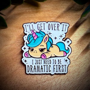 Könnte beinhalten: Ein skurriler Emaille-Pin mit einem Cartoon-Einhorn-Design. Das Einhorn hat eine türkisfarbene Mähne und ein Horn und einen mürrischen Gesichtsausdruck. Der Pin trägt den Text "I'll get over it, I just need to be dramatic first."