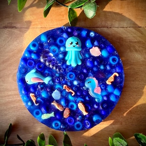 Op de afbeelding: Een ronde blauwe hars onderzetter ingelegd met diverse oceaan-thema bedels, waaronder een lichtblauwe kwal, een blauw zeepaardje, een blauwe dolfijn, verschillende kleine vissen en kleine schelpjes, omringd door talloze doorzichtige blauwe kralen.