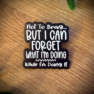 Könnte beinhalten: Ein schwarzer Emaille-Pin mit weißem Text, der lautet: Not To Brag... BUT I CAN FORGET WHAT I'M DOING While I'm Doing It. Der Pin hat eine gestanzte Form, die dem Umriss der Schrift folgt, und zeichnet sich durch eine glatte Oberfläche aus.