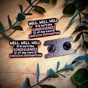 Puede incluir: Dos pines de esmalte con el texto "WELL, WELL, WELL if it isn't the CONSEQUENCES of my own EXECUTIVE DYSFUNCTION" en negro, rosa y blanco. Los pines est&aacute;n sobre una superficie de madera con hojas verdes.