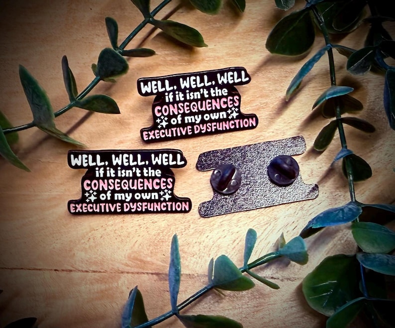 Puede incluir: Dos pines de esmalte con el texto "WELL, WELL, WELL if it isn't the CONSEQUENCES of my own EXECUTIVE DYSFUNCTION" en negro, rosa y blanco. Los pines est&aacute;n sobre una superficie de madera con hojas verdes.