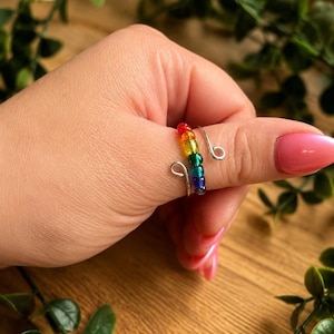 Könnte beinhalten: Ein silberfarbener Drahtring mit einer Reihe kleiner, durchscheinender Perlen in einer Regenbogenfarbfolge aus Rot, Orange, Gelb, Grün, Blau und Violett. Der Ring hat ein offenes, verstellbares Design mit geschlungenen Enden an beiden Seiten.