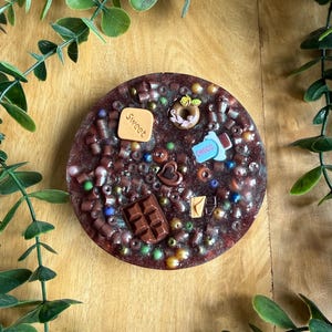 Può includere: Un sottobicchiere circolare in resina marrone scuro con incastonate varie piccole perline e ciondoli in miniatura a tema alimentare, tra cui un quadrato color sabbia con la scritta Sweet, una ciambella marrone, un pezzo blu con la scritta Choco, un cuore e un pezzetto di cioccolato.