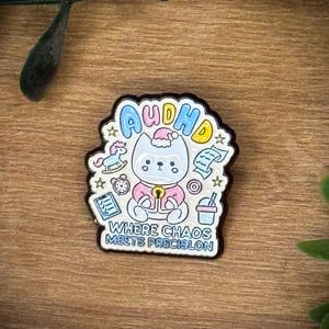 Peut inclure: Une broche en émail métallique représentant un chat blanc portant un pull rose et un bonnet de Père Noël. Le texte indique "AuDHD" en haut et "WHERE CHAOS MEETS PRECISLON" en bas, entouré de petites icônes de cheval à bascule et d'horloge.
