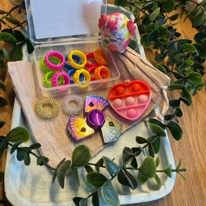 Könnte beinhalten: Eine Sammlung von sensorischen Spielzeugen, darunter bunte Ringe, eine Axolotl-Figur mit Batikmuster, ein herzförmiges Pop-it und ein Fidget Spinner. Die Artikel sind auf einem Tablett mit einem beigefarbenen Beutel und Grünzeug angeordnet.