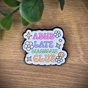 Könnte beinhalten: Ein weißer Emaille-Pin mit schwarzem Rand, der den Text ADHD LATE DIAGNOSIS CLUB in bunten Buchstaben zeigt. Das Design enthält dekorative Elemente wie orangefarbene und gelbe Sterne, weiße Blumen mit orangefarbenen Mitten und ein kleines Smiley-Gesicht.