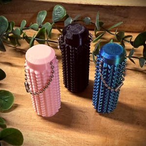 Può includere: Tre tappi per bottiglie decorativi e strutturati in rosa, nero e blu. Ogni tappo ha una forma cilindrica con un motivo a coste e una piccola catena attaccata. I tappi sono disposti su una superficie di legno con fogliame verde.