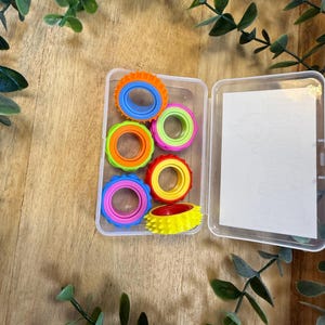 Könnte beinhalten: Eine transparente Plastikbox enthält sechs bunte Zappelringe. Die Ringe sind in den Farbtönen Orange, Blau, Grün, Rosa und Gelb gehalten. Die Box ist geöffnet und zeigt einen leeren weißen Raum. Der Hintergrund ist eine Holzoberfläche mit grünen Blättern.