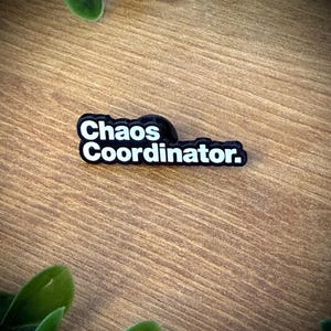 Könnte beinhalten: Ein schwarzer, rechteckiger Anstecker mit abgerundeten Kanten und dem weißen Schriftzug Chaos Coordinator. Der Anstecker hat eine kräftige serifenlose Schrift und eine glatte Oberfläche, die einen klaren Kontrast zwischen dem schwarzen Hintergrund und der weißen Schrift bildet.