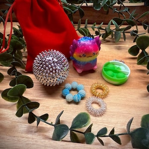 Könnte beinhalten: Eine Sammlung von sensorischem Spielzeug, darunter ein stacheliger Massageball, ein buntes, außerirdisch geformtes Spielzeug, ein grün-weißes Pop-it und mehrere Fingerringe. Im Hintergrund befindet sich ein roter Samtbeutel. Die Spielzeuge sind auf einer Holzoberfläche angeordnet.