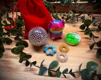 Puede incluir: Una colección de juguetes sensoriales, que incluye una bola de masaje con púas, un juguete con forma de alienígena colorido, un pop-it verde y blanco y varios anillos para los dedos. Una bolsa de terciopelo rojo está en el fondo. Los juguetes están colocados sobre una superficie de madera.