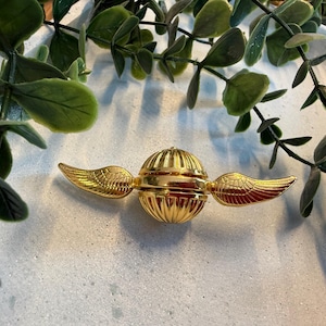 Pode incluir: Um ornamento dourado de Pomo de Ouro com asas detalhadas e um corpo esférico. O ornamento tem um design canelado e está sobre um fundo claro com folhagem verde.