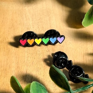 Könnte beinhalten: Zwei schwarze Metall-Emaille-Anstecknadeln, die jeweils eine Reihe von sechs kleinen Herzen in einem Regenbogenverlauf aus Rot, Orange, Gelb, Grün, Blau und Violett zeigen. Die Pins haben ein schwarzes Finish und zwei Schmetterlingsverschlüsse.