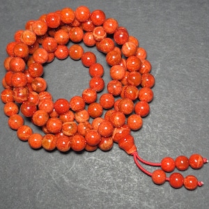 May include: A strand of orange-red beads, likely coral, arranged in a circular pattern. The beads are round and have a natural, textured appearance. A tassel of smaller beads and red string hangs from the strand. The beads are on a dark gray surface.