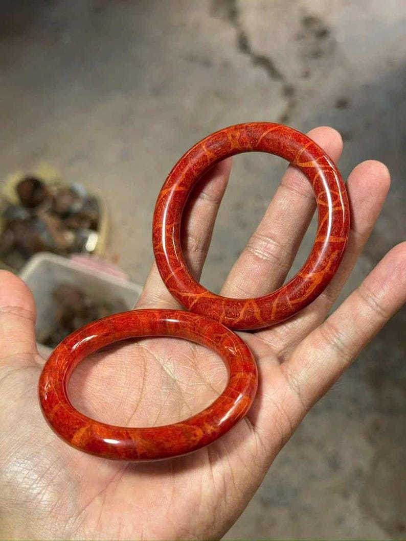 May include: Two red coral bangles with a natural, textured pattern. The bangles are round and polished, showcasing a vibrant red hue with lighter orange veins. The jewelry is displayed on a hand.